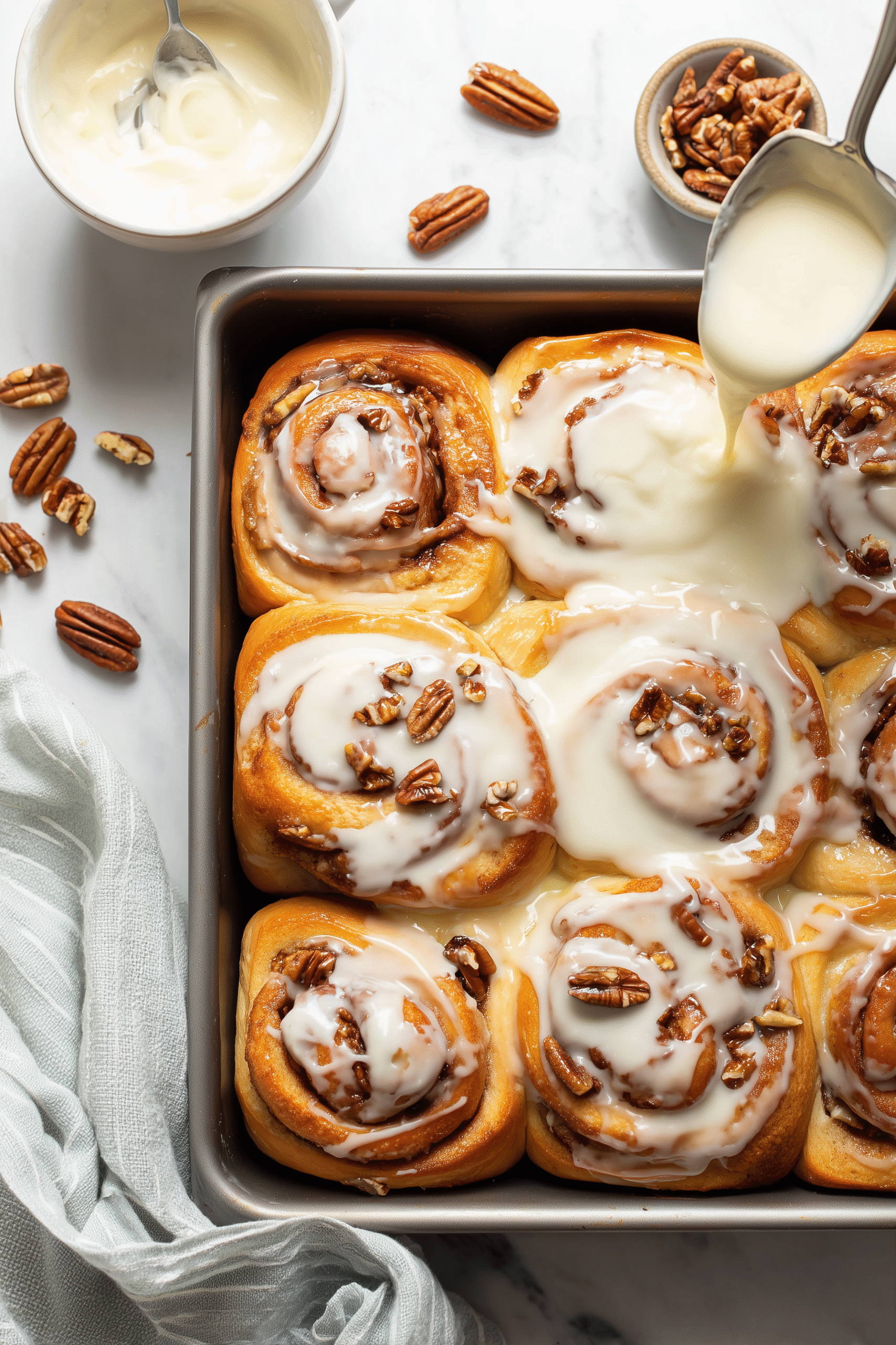 Baked homemade cinnamon rolls being iced in a pan on white marble.
