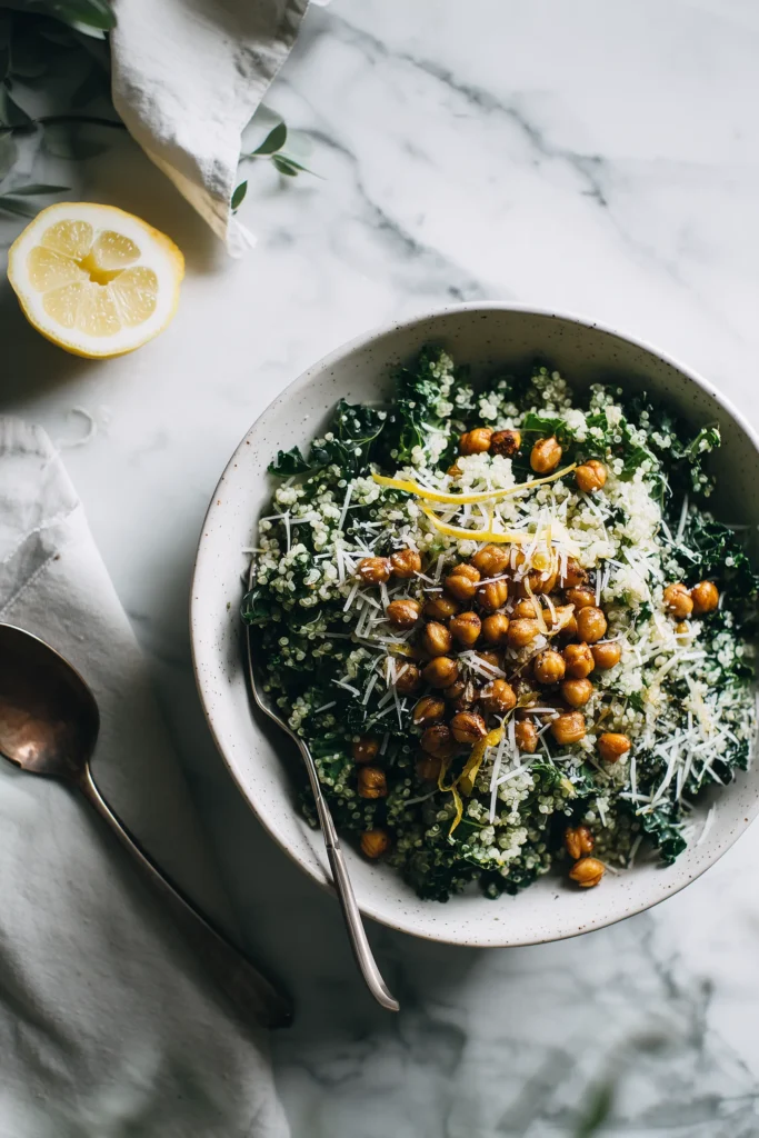 A wide bowl with Lemony Quinoa Kale Salad, garnished with roasted chickpeas and Parmesan on a marble surface.