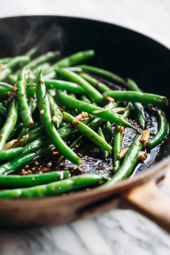 Green beans stir-fried in a skillet with garlic sauce, showing steam and a glossy finish.