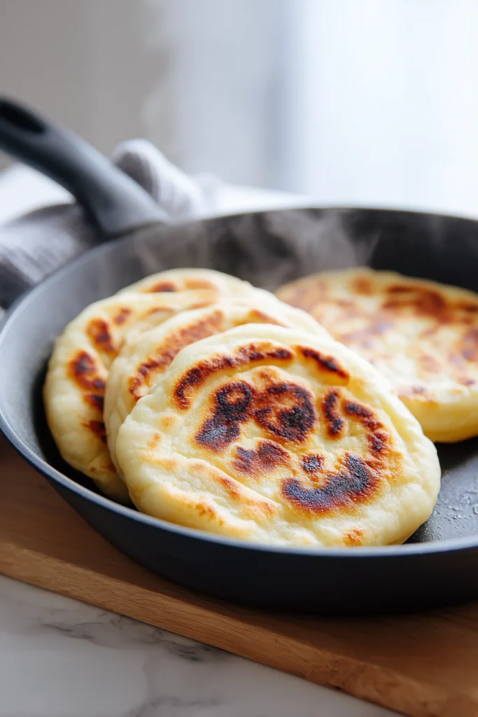 Several pieces of Cottage Cheese Flatbread (3 Ingredients) sizzling in a skillet, golden and pillowy on a light wood background.