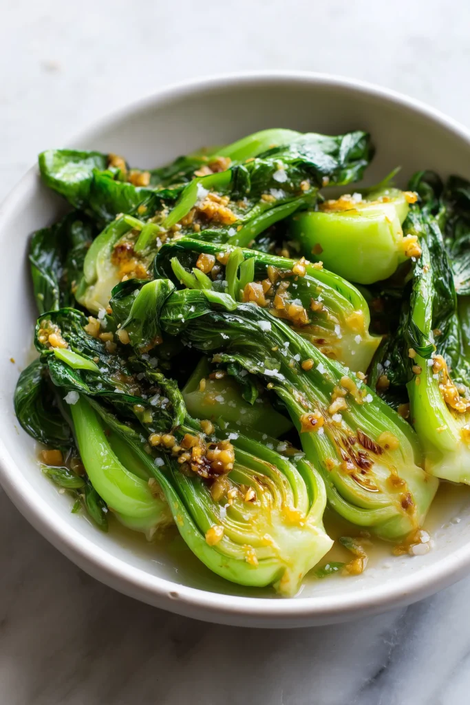 A bright modern close-up of Garlic Ginger Bok Choy in a white bowl showing glossy greens, caramelized edges, and bits of garlic and ginger.
