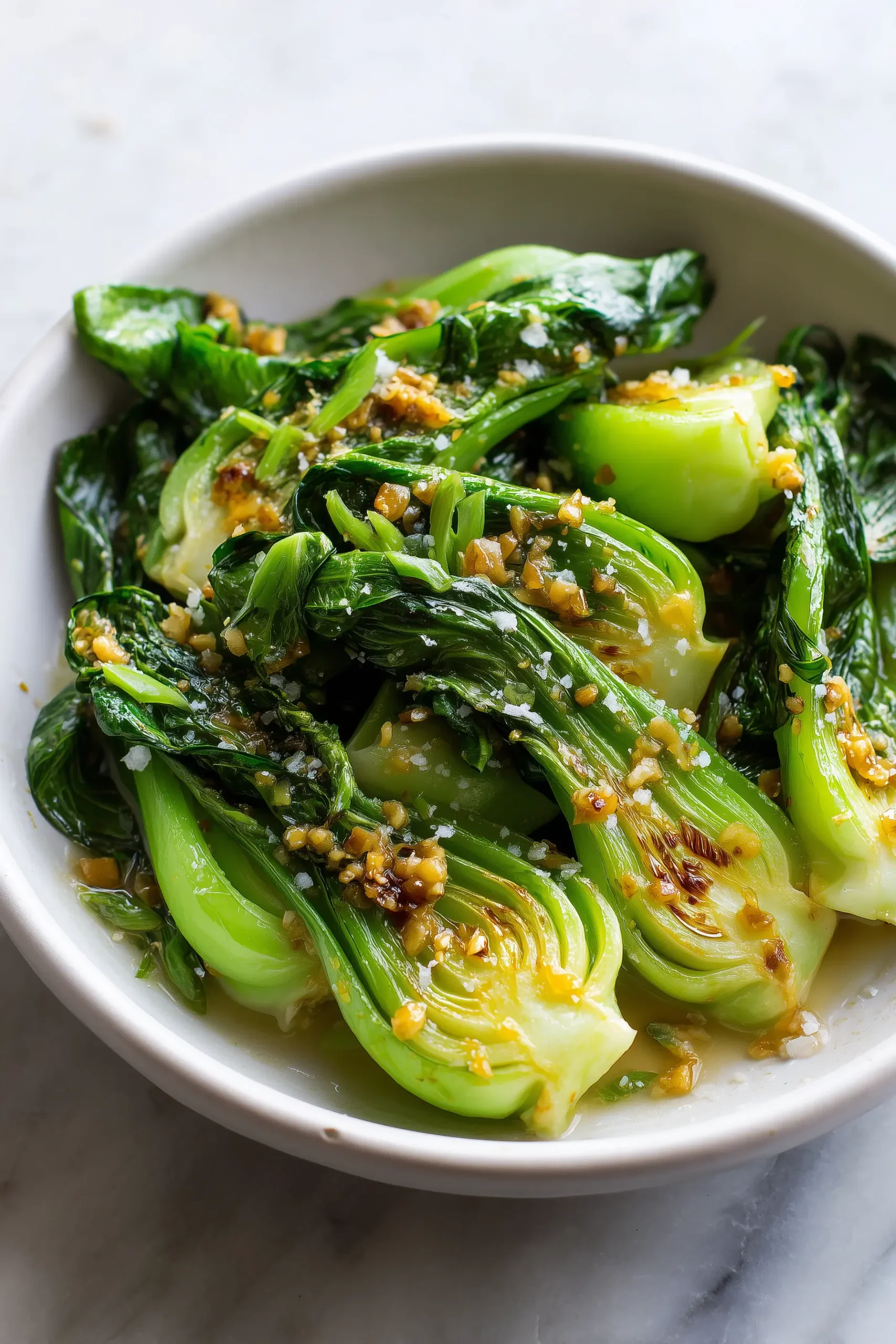A bright modern close-up of Garlic Ginger Bok Choy in a white bowl showing glossy greens, caramelized edges, and bits of garlic and ginger.