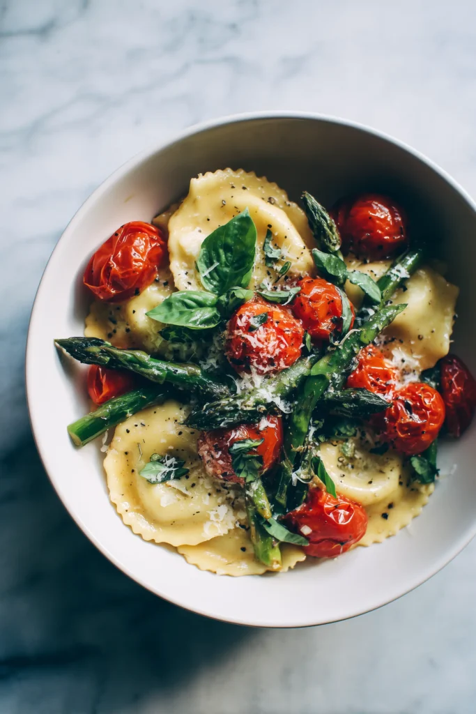 A bowl of Ravioli with Tomatoes and Asparagus tossed in olive oil and herbs sits on a bright marble surface.