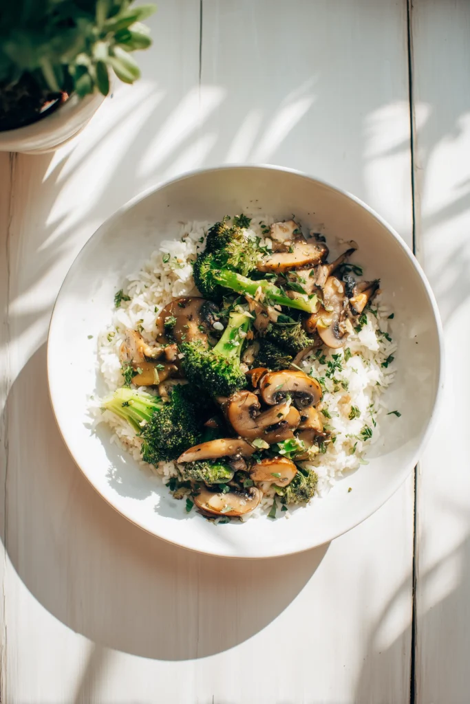 Sauteed mushrooms with broccoli served atop white rice, garnished with herbs on a clean white plate.