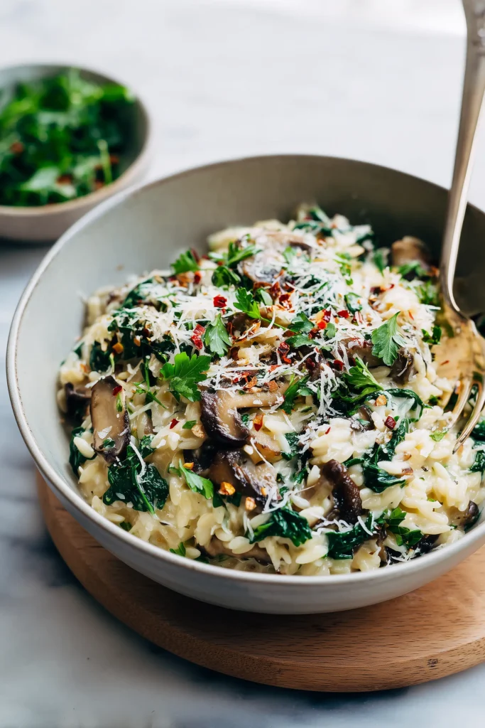 Spinach Mushroom Orzo 4 A bowl of Spinach Mushroom Orzo, garnished with cheese and parsley, next to a salad on a light wood table.