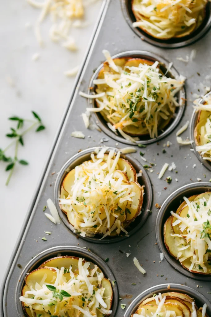 Crispy Cheddar Potato Stacks with Lacy Edges & Creamy Centers 3 Close-up of muffin tin cups filled with layered potato slices, cheddar cheese, and herbs for buttsharp cheddar potato stacks on a white marble surface.