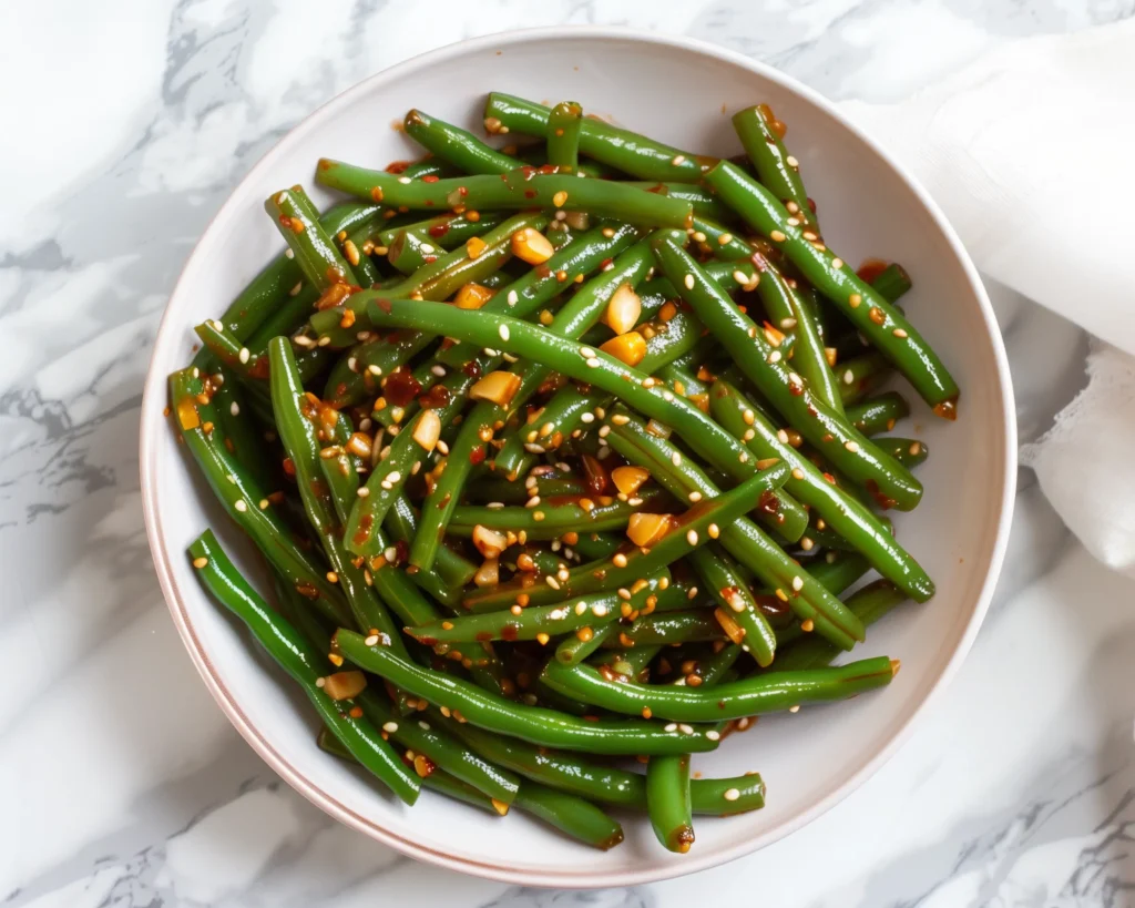 A bowl of garlic chili green beans tossed with chili oil and sesame seeds, bright and fresh on white marble.