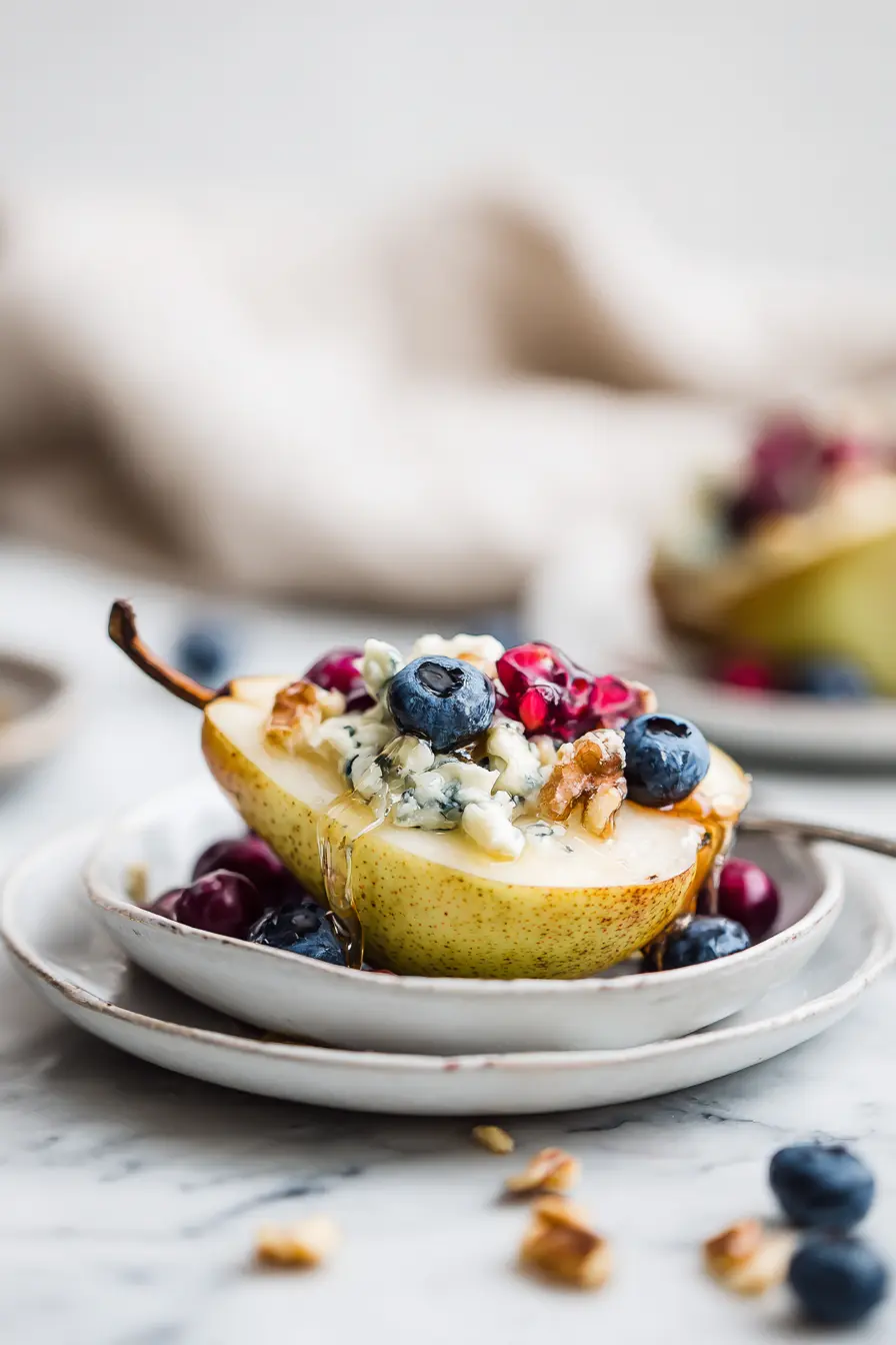 Baked Pears with Berries, Honey, Walnuts & Blue Cheese (Cozy, Easy Dessert) 4 Single serving of Baked Pears with Berries, Honey, Walnuts & Blue Cheese on a white plate over white marble, topped with berries, walnuts, and melted blue cheese.